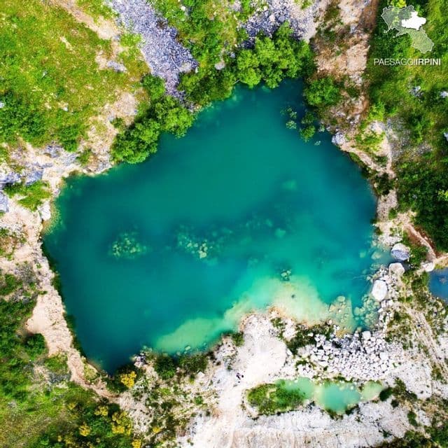 Una vista aerea, dall'alto, di un lago turchese circondato da sponde rocciose ed erbose e alberi verdi.
