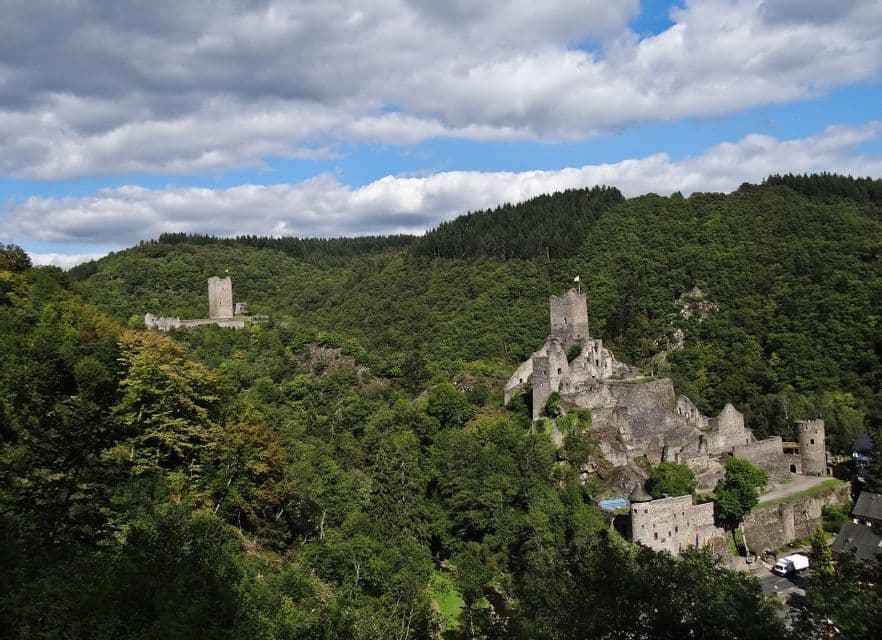 Due rovine di castelli in pietra si ergono su colline opposte coperte da una fitta foresta verde sotto un cielo azzurro nuvoloso.