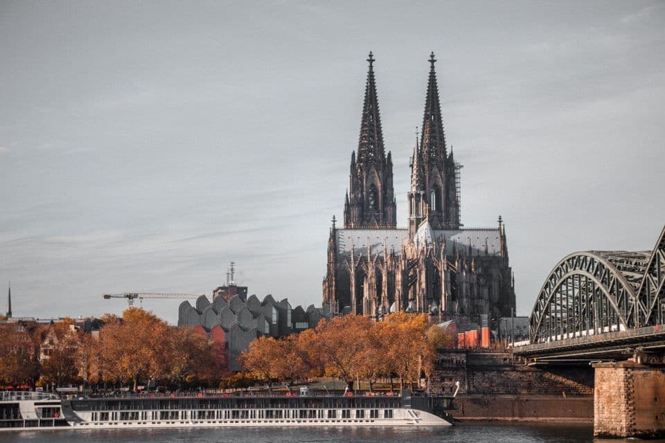 Un paesaggio urbano con una grande cattedrale gotica e un ponte ad arco visti da oltre un fiume, con alberi autunnali arancioni in primo piano.