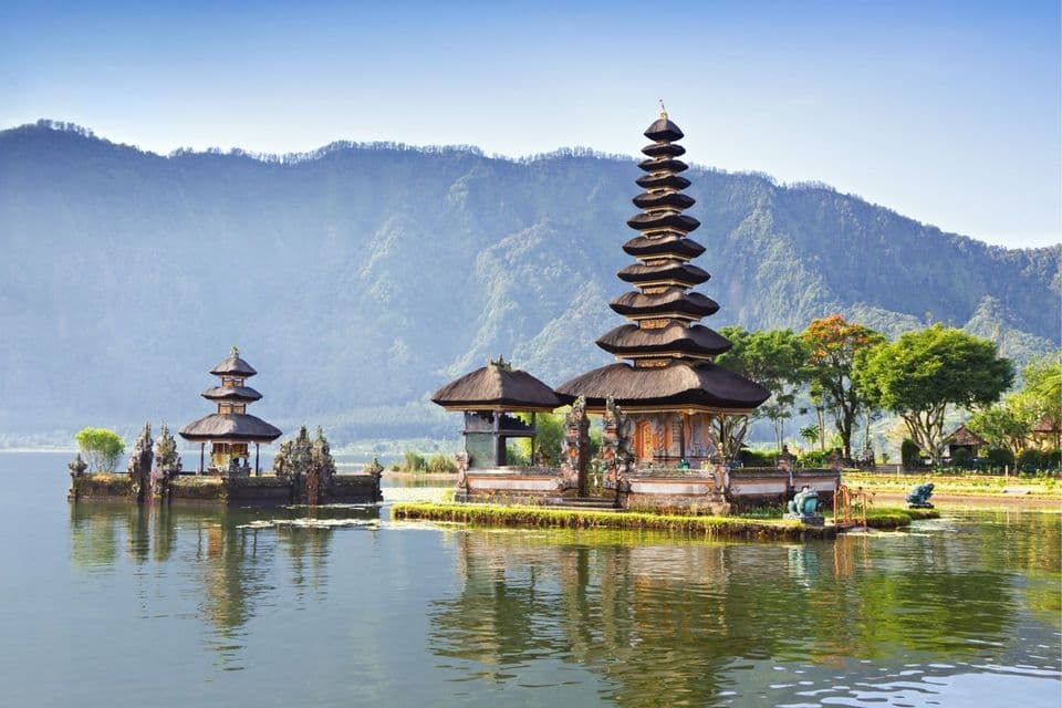 Ein traditioneller Tempelkomplex im Pagodenstil liegt an einem ruhigen See, mit einem bewaldeten Gebirgszug im Hintergrund.