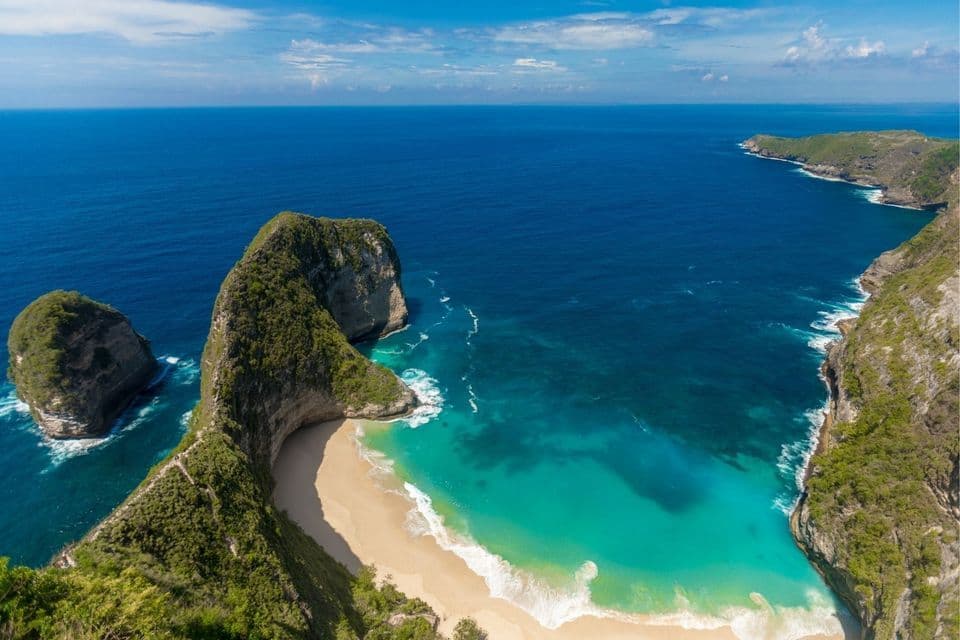 Luftaufnahme einer grünen, felsigen Küste mit einer markanten Halbinsel, die einen weißen Sandstrand und türkisfarbenes Wasser schützt.
