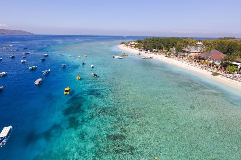 Luftaufnahme einer tropischen Küste mit vielen kleinen Booten in klarem türkisfarbenem Wasser neben einem weißen Sandstrand mit Bäumen und Gebäuden.