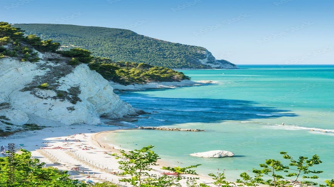 Una spiaggia soleggiata con ombrelloni incastonata tra scogliere bianche e acqua turchese, con una collina boscosa sullo sfondo.
