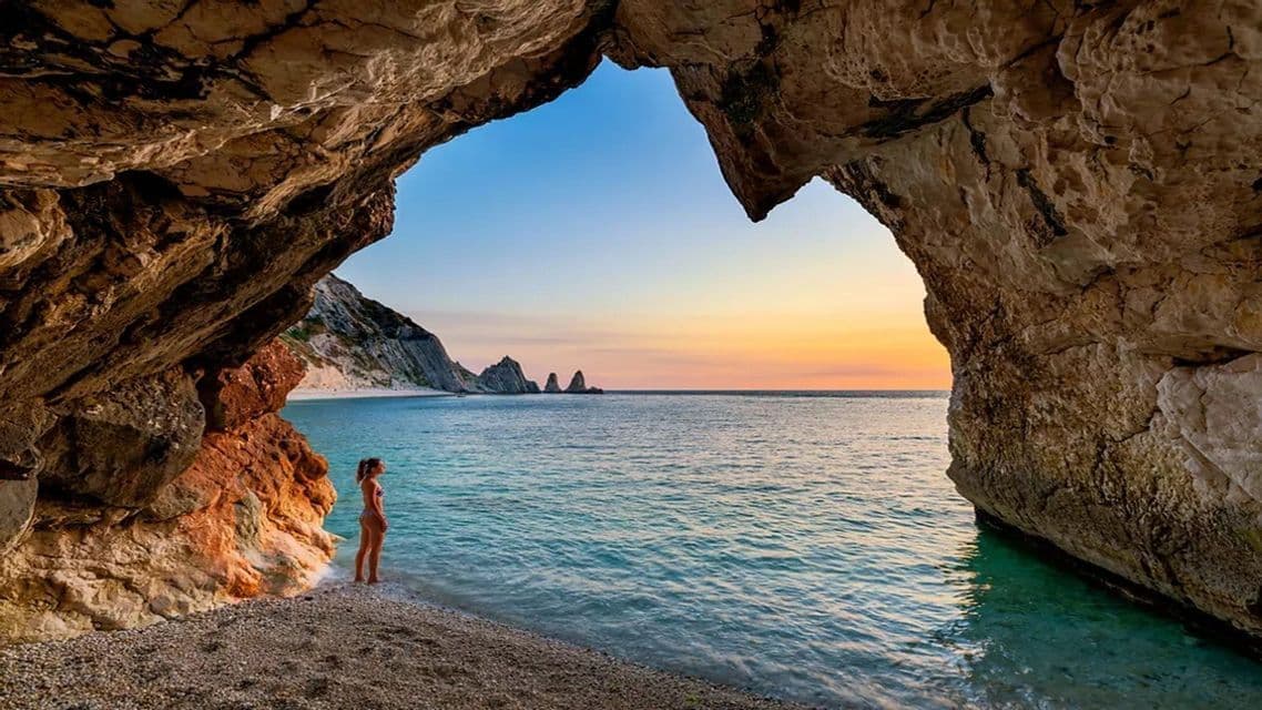 Una donna in costume da bagno si trova su una spiaggia di ciottoli all'interno di un'ampia grotta rocciosa, guardando un mare calmo con faraglioni al tramonto.