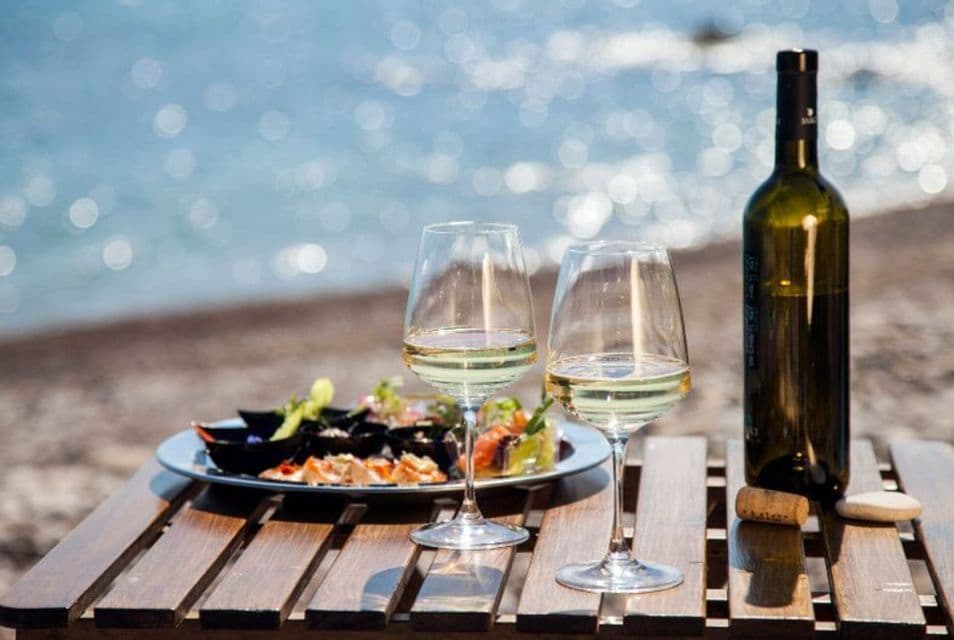 Due calici di vino bianco, una bottiglia e un piatto di antipasti si trovano su un tavolo di legno in spiaggia con il mare sullo sfondo.