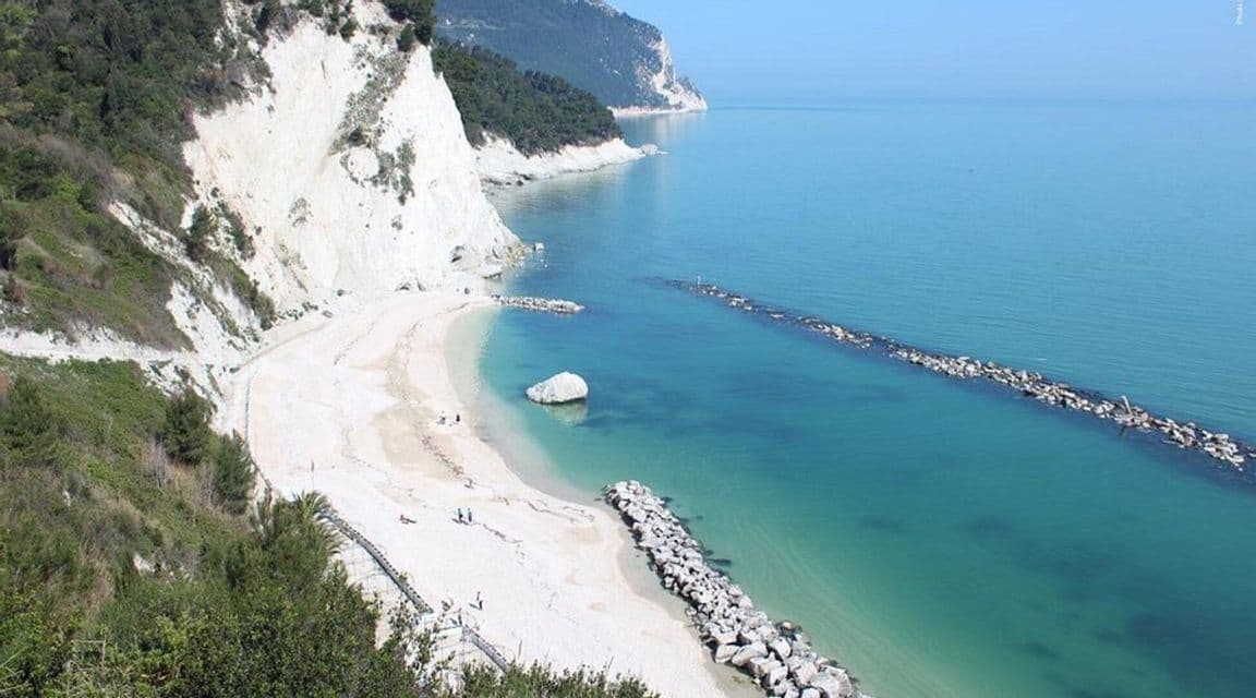 Vista dall'alto di una spiaggia sabbiosa appartata ai piedi di scogliere bianche, con acqua turchese e frangiflutti di roccia.