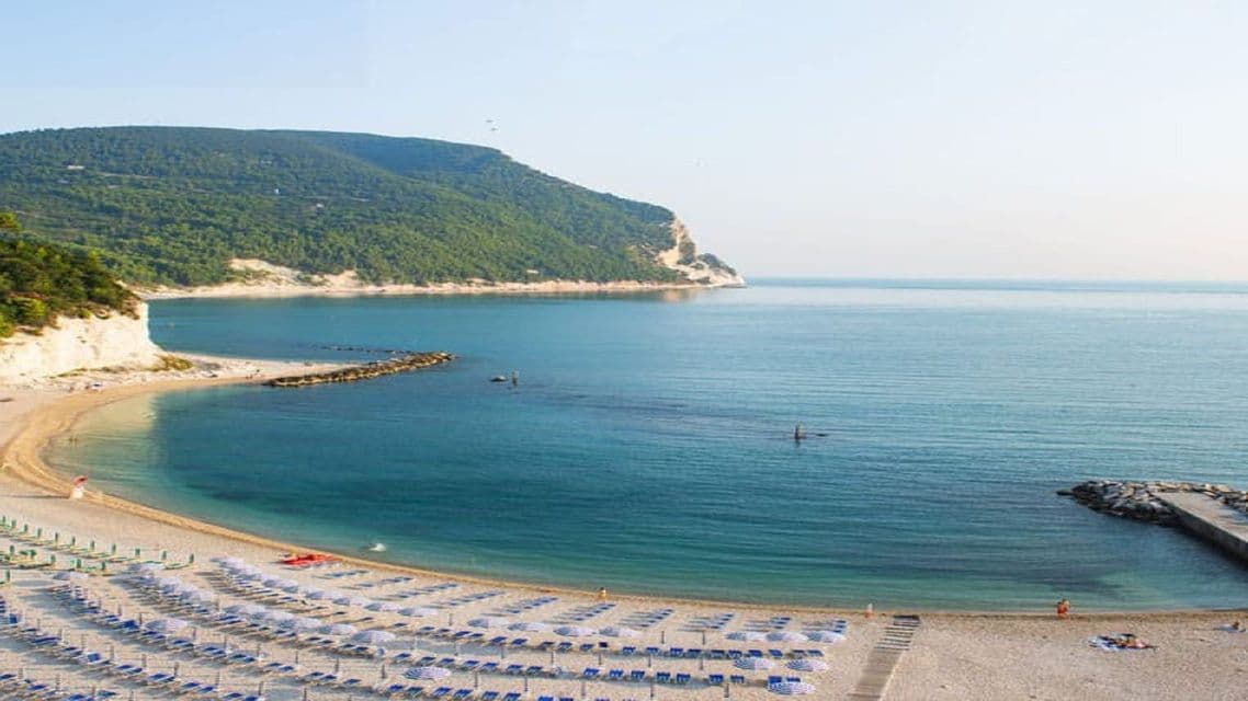 Cala di sabbia con lettini e ombrelloni ben disposti, accanto a mare calmo e blu e una grande collina verde.