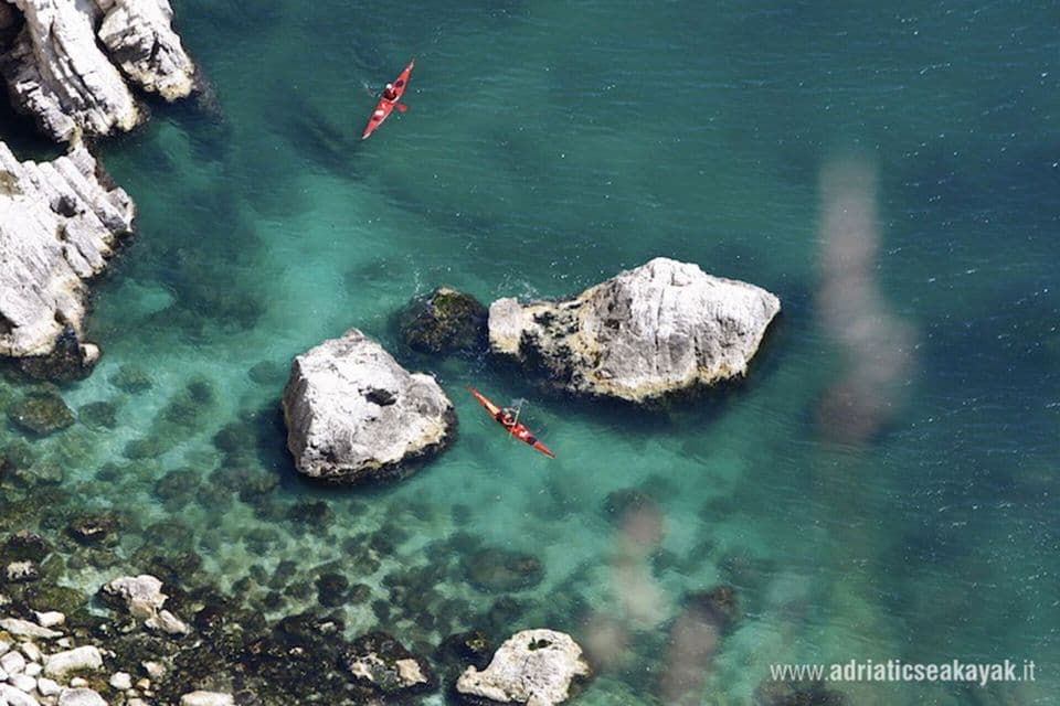Vista aerea di due persone in kayak rossi durante un viaggio di gruppo WeRoad, che remano in acque turchesi e limpide vicino a una costa rocciosa.