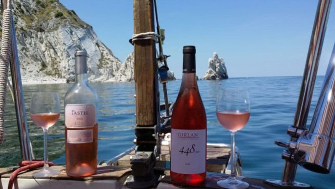 Due bottiglie e due bicchieri di vino rosato sul ponte di una barca a vela, con scogliere rocciose sullo sfondo.