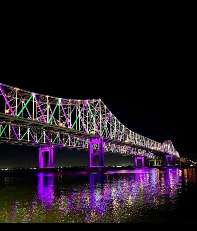 A large truss bridge illuminated with purple and green lights at night, its colors reflecting on the water below.