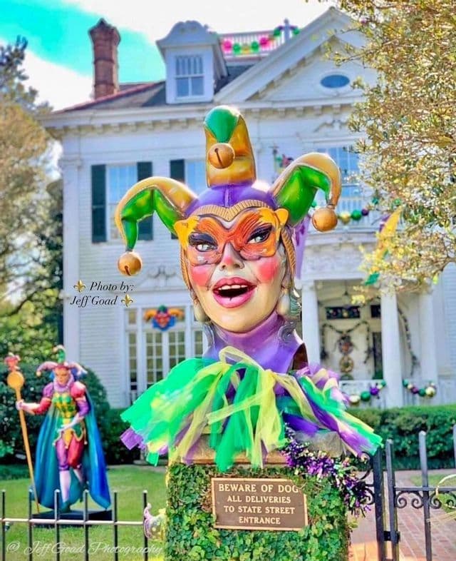 A large, colorful Mardi Gras jester bust sits on a pedestal in front of a white two-story house.