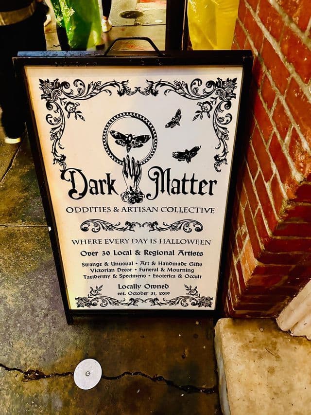 A black and white A-frame sign for 'Dark Matter Oddities & Artisan Collective' with gothic text and moth illustrations on a sidewalk.