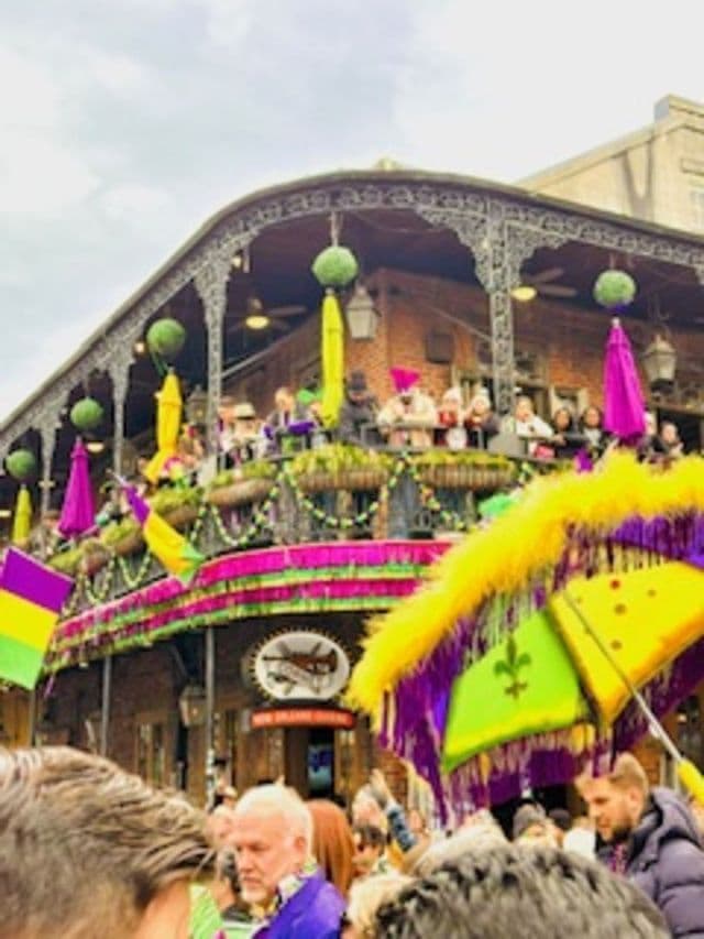 A festive crowd celebrates on a street during a carnival, with people on a decorated balcony.