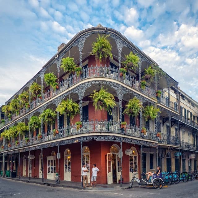 A red brick corner building with ornate metal balconies covered in hanging green ferns under a blue sky.