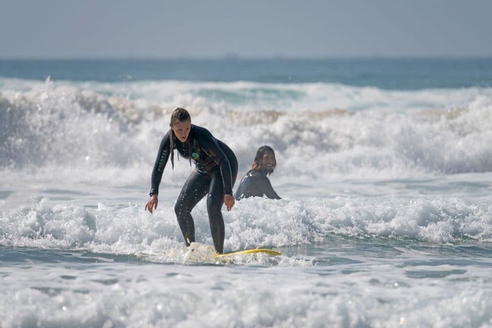 A woman from a WeRoad group trip stands on a yellow surfboard, riding a wave in the ocean.