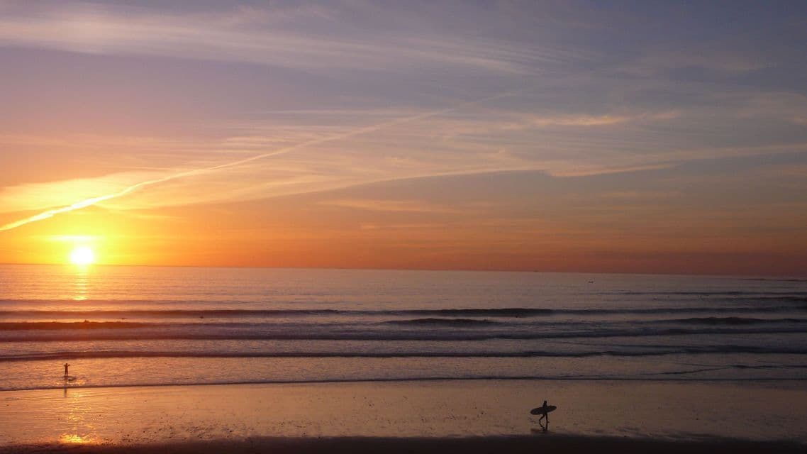 A surfer in silhouette walks along the shoreline carrying a surfboard as the sun sets over the ocean.