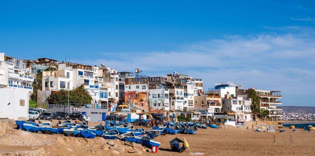 A row of blue fishing boats sits on a sandy beach below a town of whitewashed buildings on a sunny day.