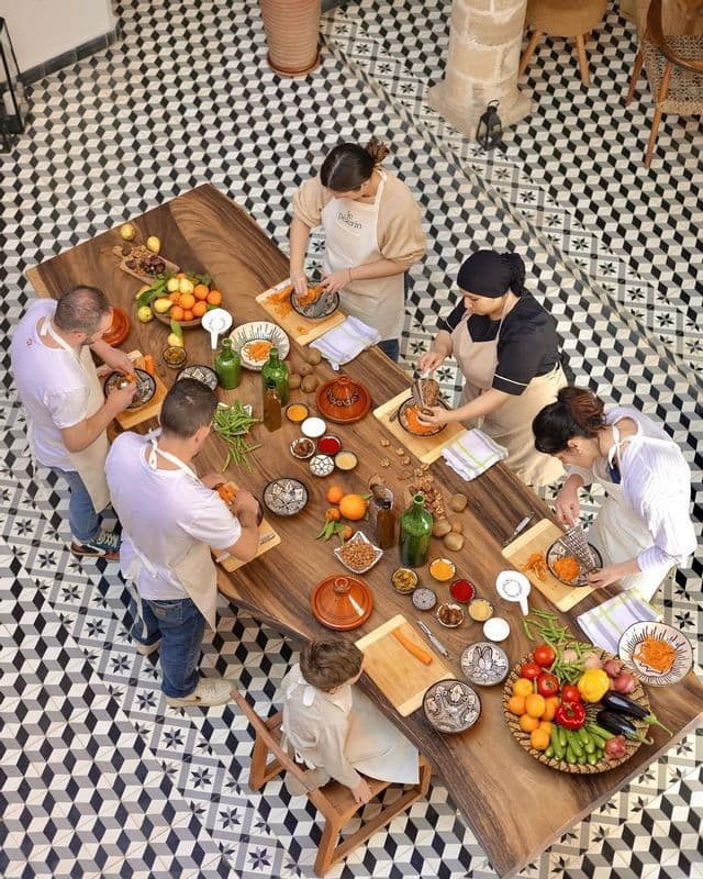 A WeRoad group trip participates in a cooking class, preparing ingredients on a wooden table with tagines, viewed from above.