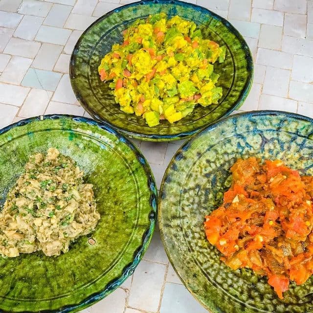 Three rustic green ceramic bowls filled with various colorful salads are arranged on a white tiled surface.