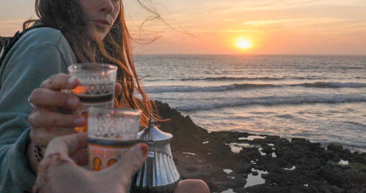Two people on a WeRoad group trip toasting with tea glasses on a rocky coast, watching the sunset over the ocean.