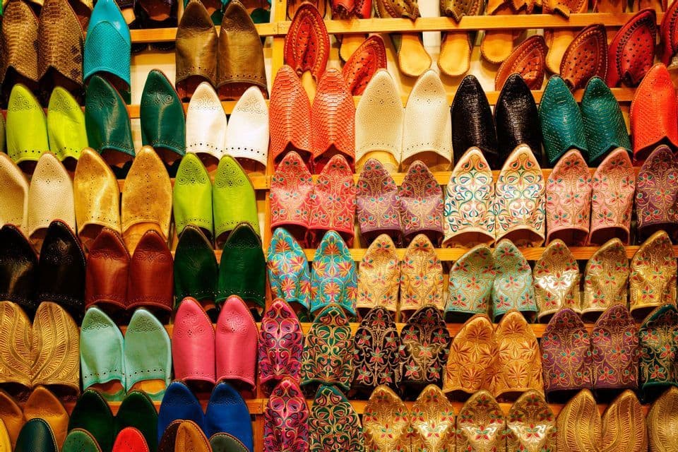A large collection of colorful, traditional Moroccan slippers, known as babouches, displayed in neat rows on wooden shelves.