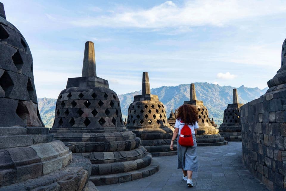 A woman with curly hair and a red backpack walks on a stone path among large, bell-shaped stupas, with mountains in the distance.