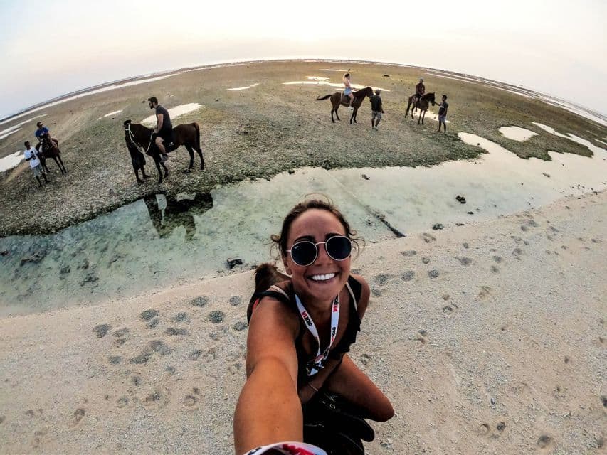 Una mujer sonriente se toma una selfie en una playa de arena mientras, en el fondo, su grupo de WeRoad cabalga a caballo durante la marea baja.
