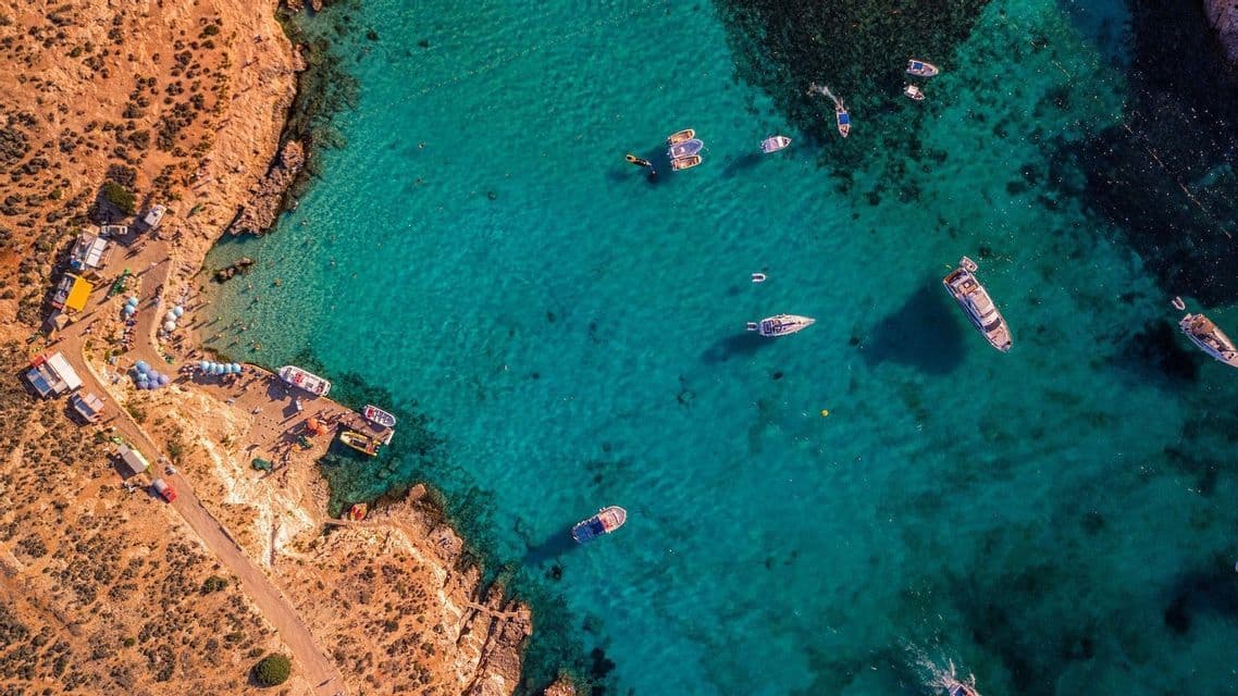 Una vista aérea, desde arriba, de una cala rocosa donde varios barcos flotan en aguas claras y turquesas junto a una pequeña playa.