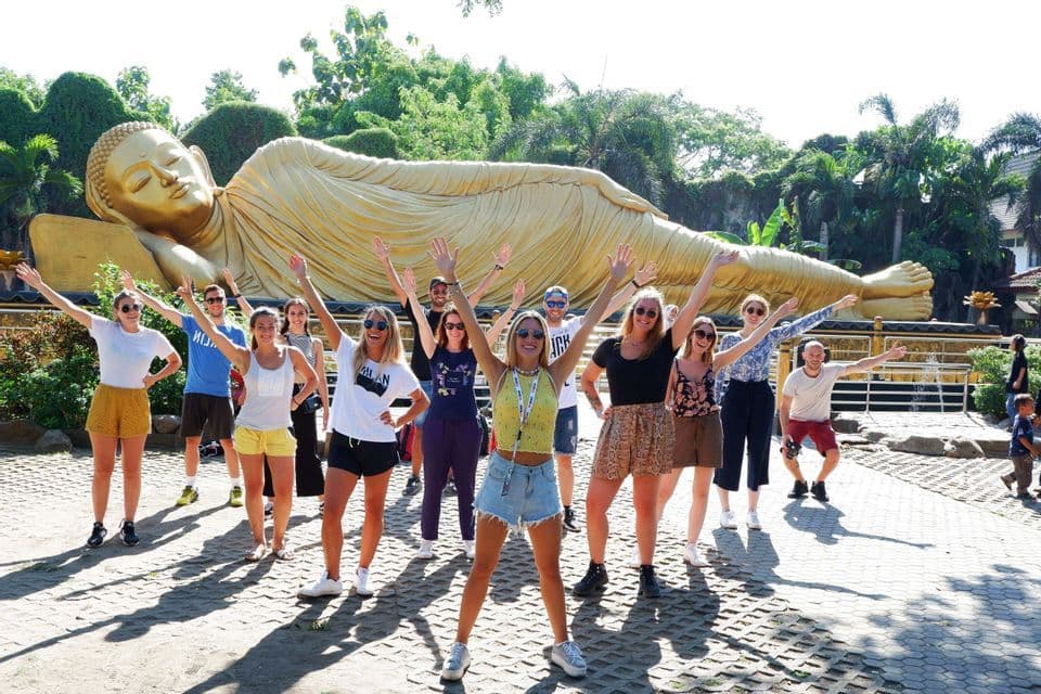 Un viaje en grupo de WeRoad posando con los brazos en alto frente a una estatua gigante y dorada de Buda reclinado.