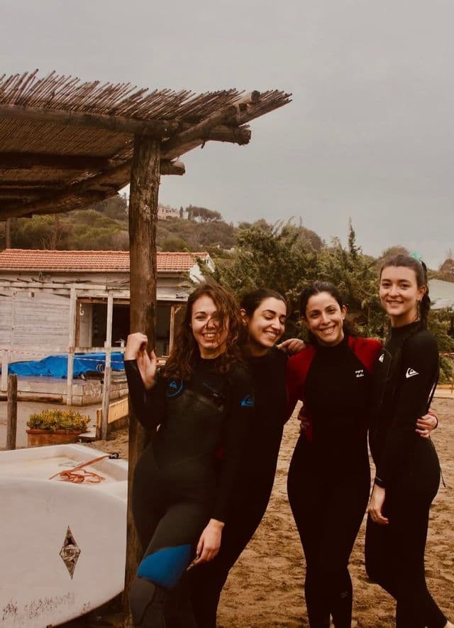 Un viaggio di gruppo WeRoad con quattro donne in muta che sorridono insieme su una spiaggia sabbiosa accanto a un rifugio di legno.