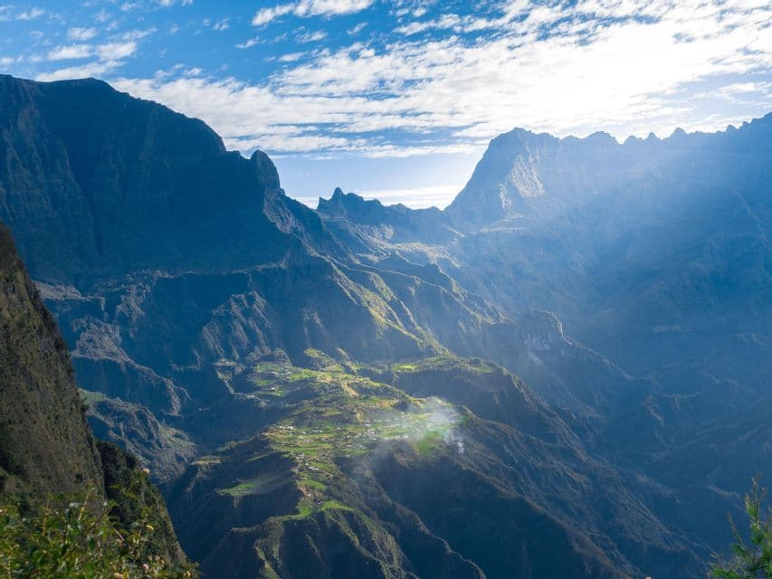 Ein kleines Dorf mit grünen Feldern liegt in einem sonnigen Tal, umgeben von massiven, schattigen Bergen unter einem blauen Himmel mit Wolken.