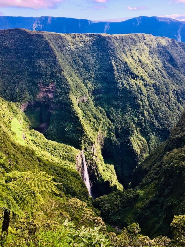 Eine Draufsicht auf einen Wasserfall, der in eine tiefe, grüne Schlucht stürzt, umgeben von üppigen Bergen unter blauem Himmel.