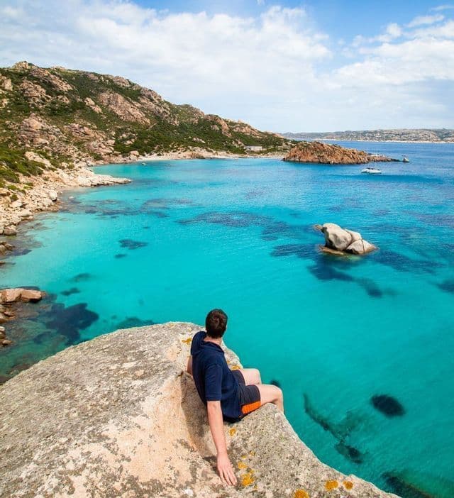 Un uomo siede su un'alta roccia che domina una baia con acque turchesi vibranti e una costa rocciosa sotto un cielo blu.