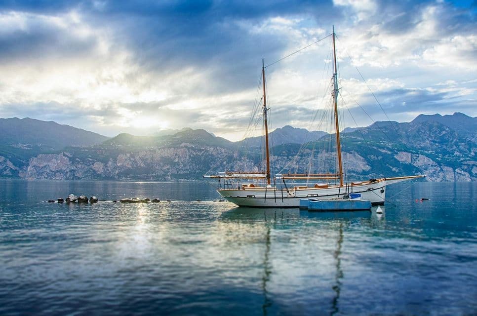 A two-masted sailboat is anchored on a calm lake with a mountain range in the background under a sunlit, cloudy sky.