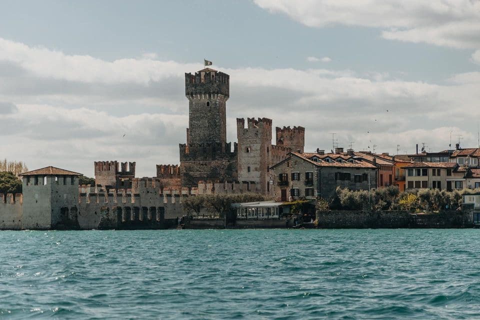 A medieval stone castle with fortified walls and towers stands on the shore of a body of water under a partly cloudy sky.