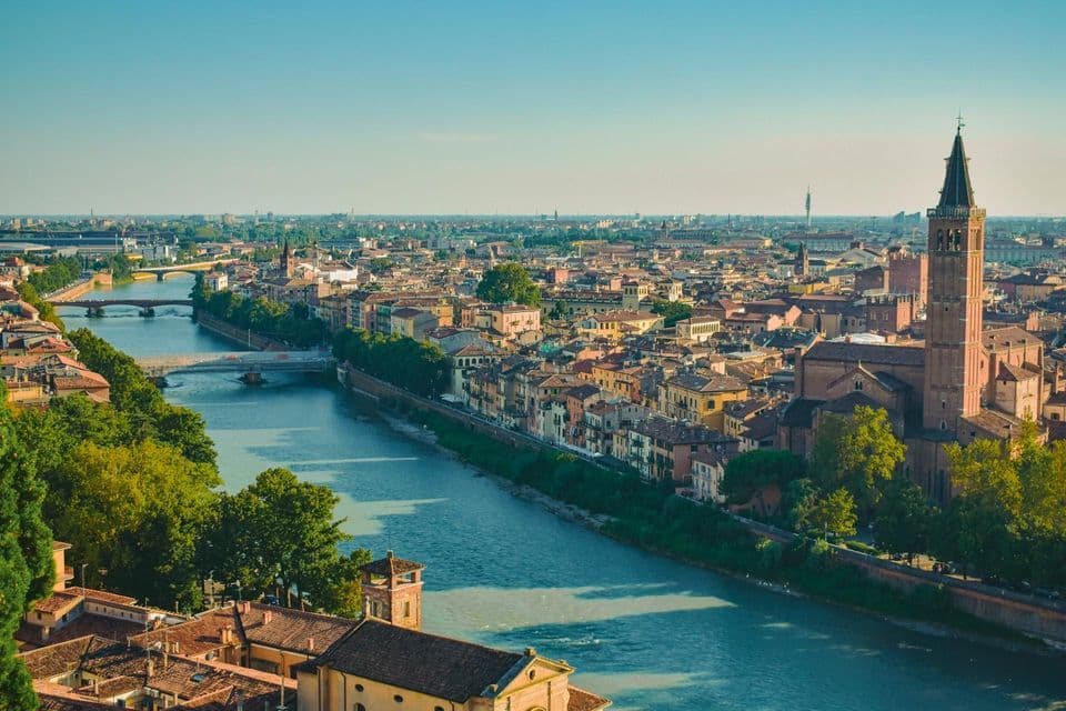 An aerial view of a historic city with a wide river, multiple bridges, terracotta roofs, and a prominent bell tower.