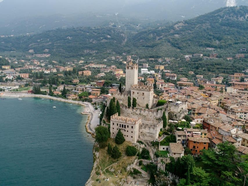 An aerial view of a stone castle on a cliff overlooking a lakeside town, with tree-covered hills in the background.