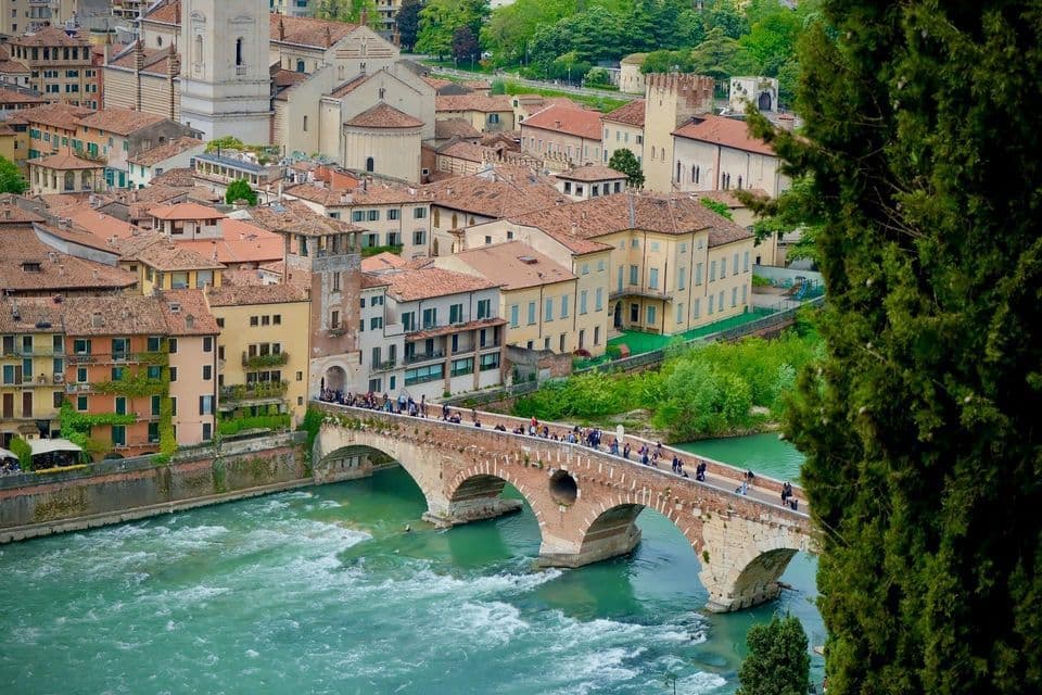 High-angle view of people crossing a stone arch bridge over a turquoise river, with historic city buildings in the background.