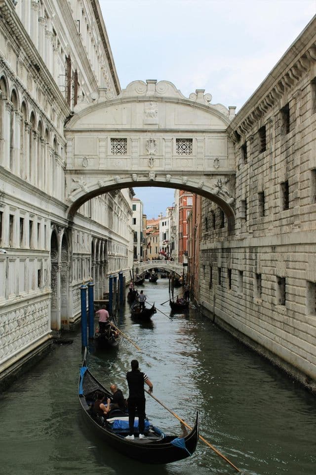 Gondoliers rowing gondolas on a narrow canal under an ornate, enclosed stone bridge connecting two historic buildings.