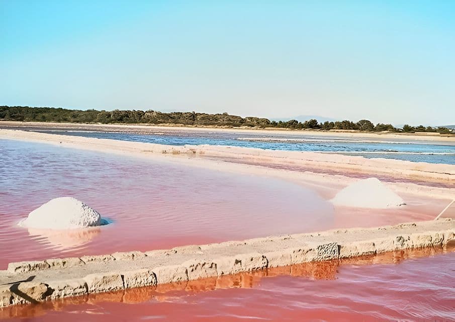 Montones de sal cosechada reposan en agua rosa en una salina bajo un cielo azul claro.