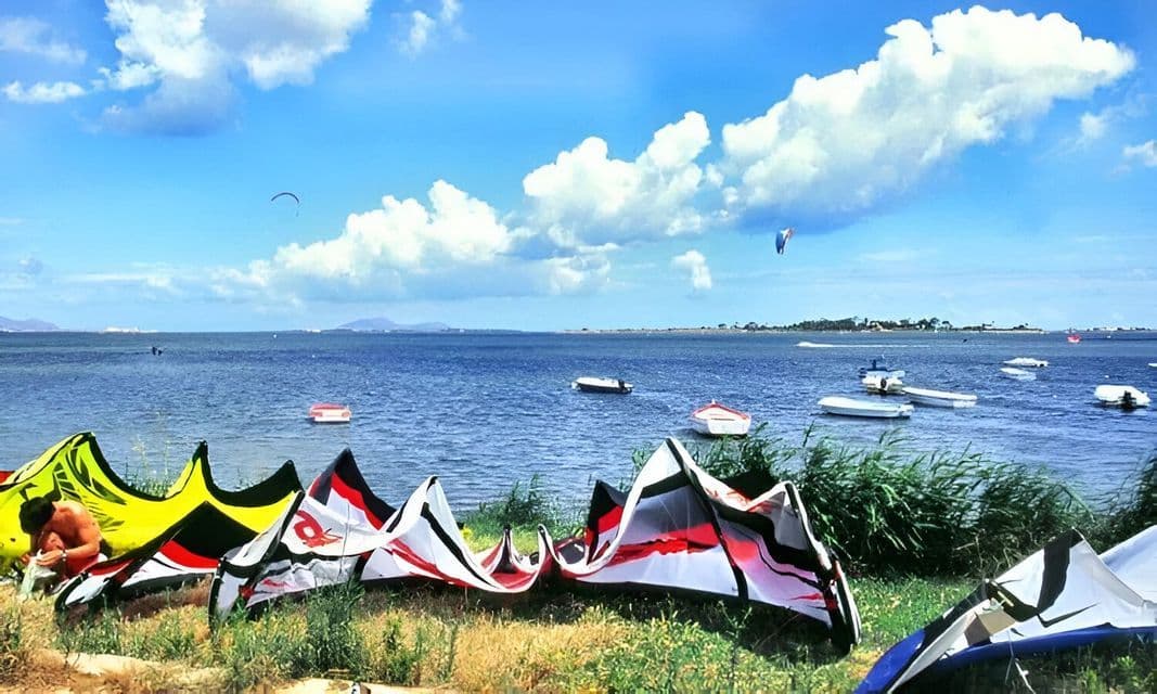 Cometas de kitesurf tendidas en una orilla cubierta de hierba junto a una bahía con varios botes pequeños bajo un cielo azul nublado.