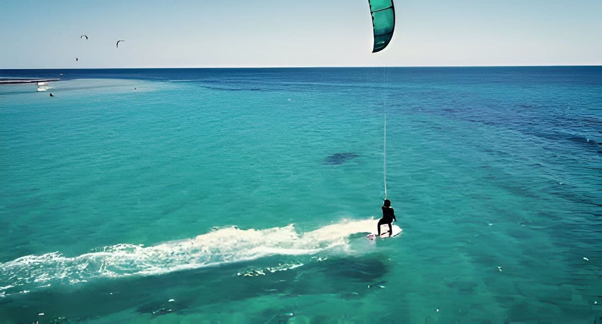 Una persona en una tabla hace kitesurf sobre agua turquesa, creando una estela bajo un cielo despejado.