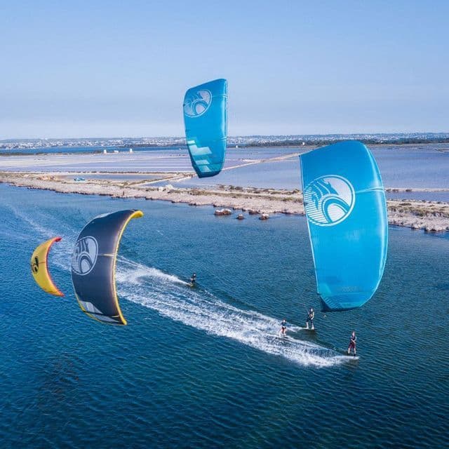 Vista aérea de un viaje en grupo de WeRoad haciendo kitesurf con cometas de colores sobre un mar azul brillante cerca de la costa.