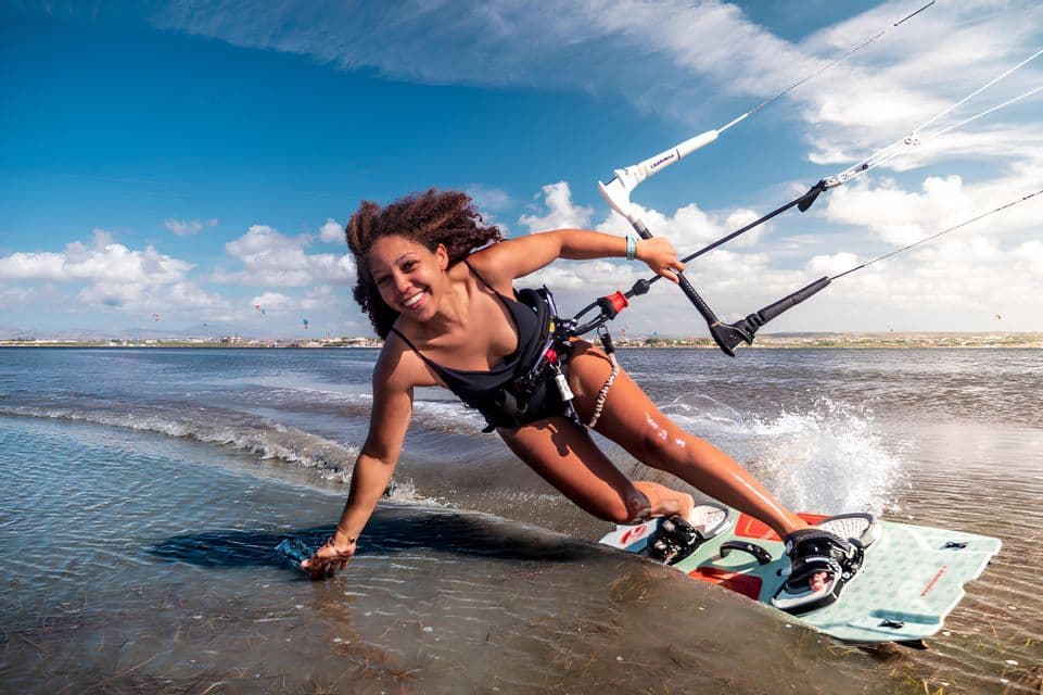 Una mujer sonriente con pelo rizado hace kitesurf en el mar, sosteniendo la barra de control bajo un cielo azul con nubes blancas.