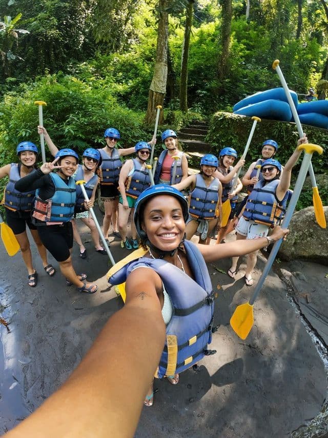 A WeRoad group trip poses for a selfie, wearing helmets and life vests and holding paddles in a lush forest.