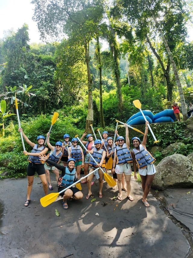 A WeRoad group trip in life vests and helmets posing with paddles on a riverbank in a lush jungle before rafting.