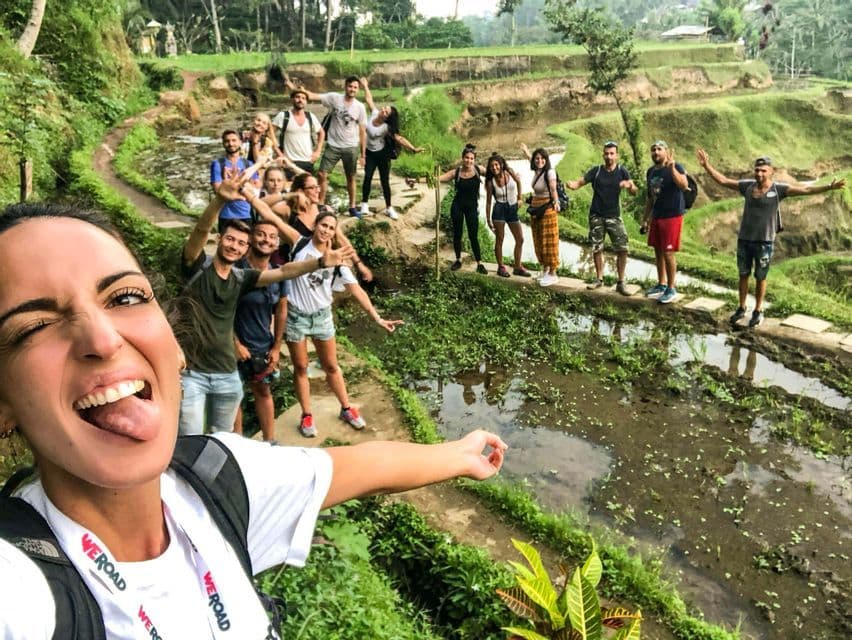 A woman takes a selfie with her WeRoad group trip as they pose on a narrow path through green terraced rice paddies.