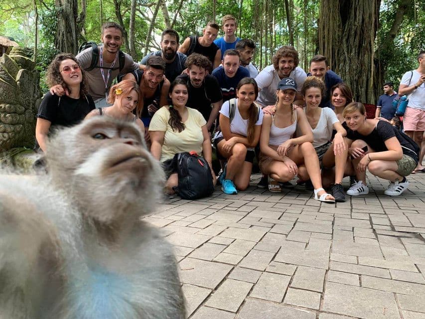A monkey takes a selfie in front of a smiling WeRoad group trip posing on a stone patio surrounded by trees.