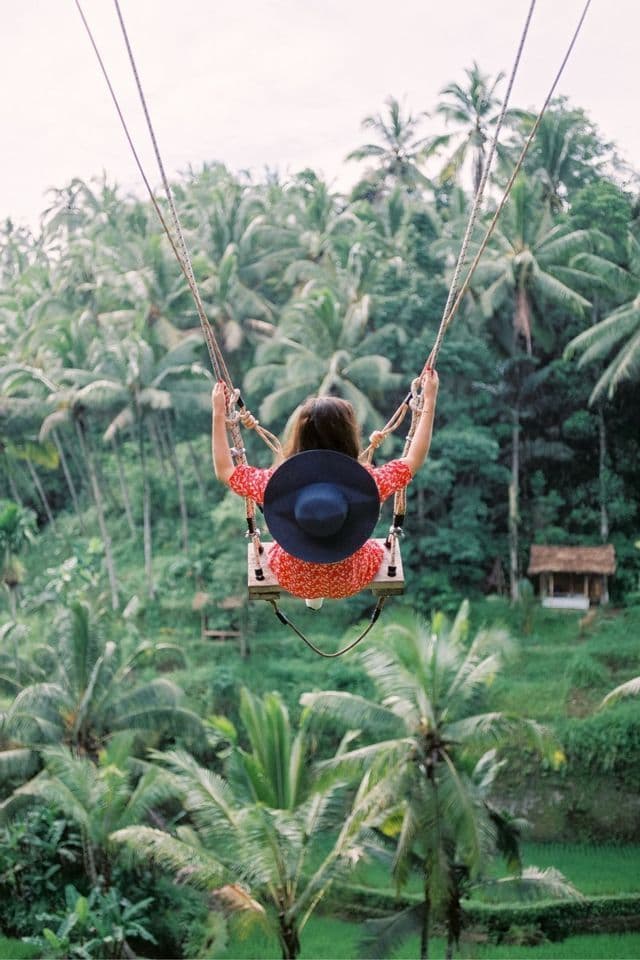 Eine Person in rotem Kleid und mit Hut schwingt auf einer großen Seilschaukel über einem üppigen Dschungel aus Palmen.
