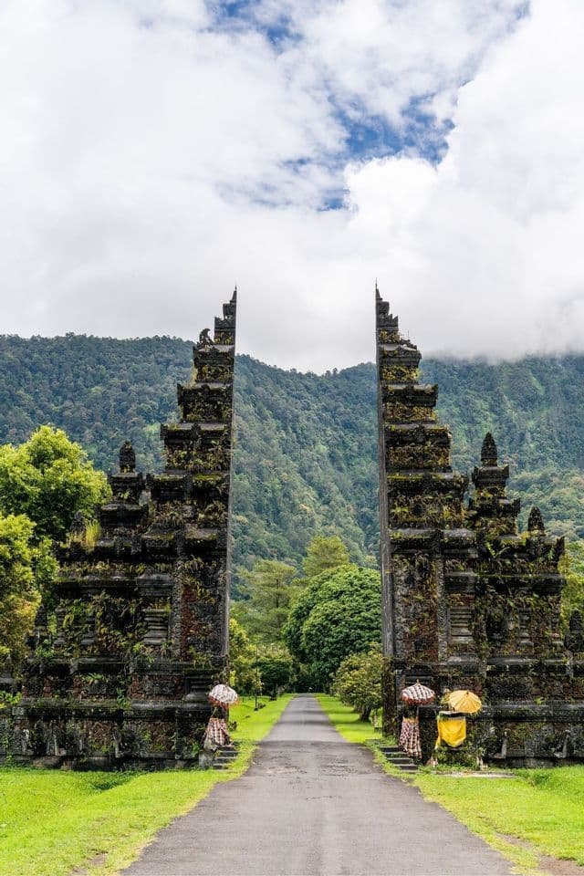 Ein hohes, verziertes Steintor, bedeckt mit Moos, rahmt einen gepflasterten Weg ein, der zu einem üppigen, bewaldeten Berg unter einem bewölkten Himmel führt.